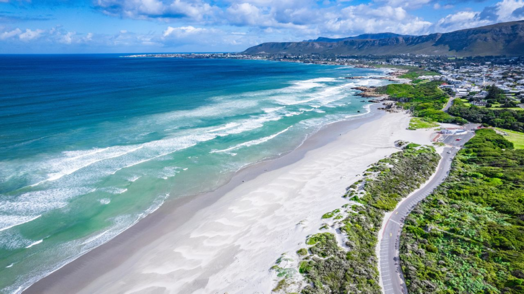 Aerial view of a long sandy beach with ocean waves, green hills, and the coastal town of Hermanus, South Africa, under a partly cloudy sky—an idyllic scene especially popular during whale season.