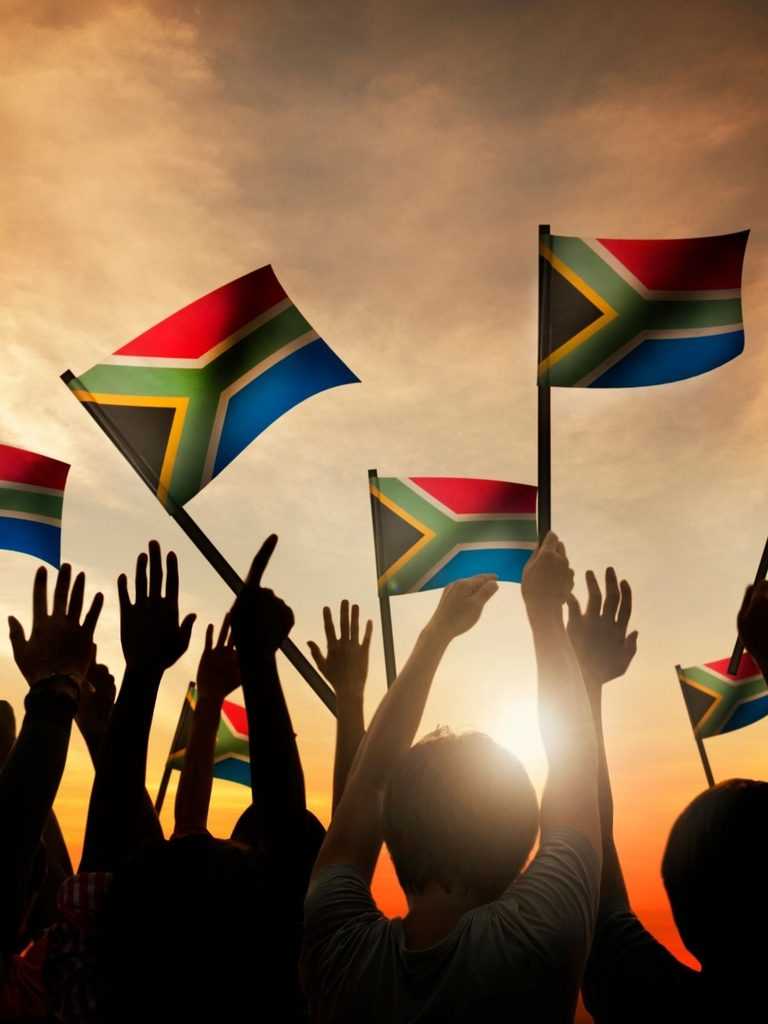 A group of people raise their hands and wave South African flags at sunset, with the sun visible in the background.
