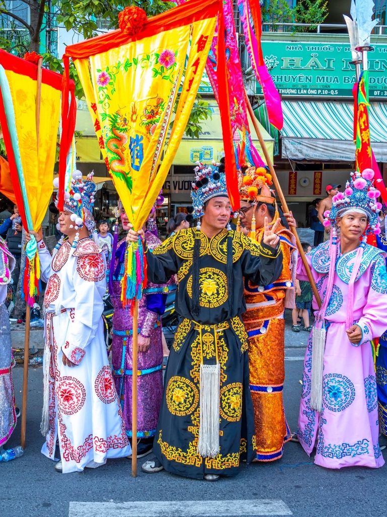 People in colorful, ornate traditional costumes stand in a street parade, holding vibrant flags and banners with decorative designs.