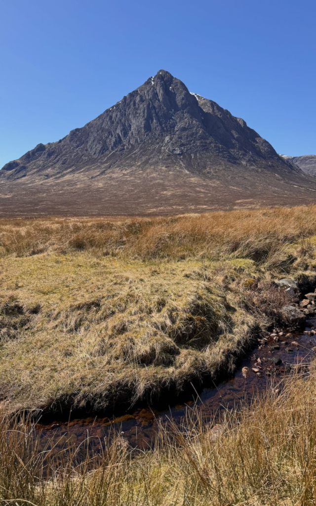 A rocky mountain rises behind a dry grassy field with a small stream running through the foreground under a clear blue sky.