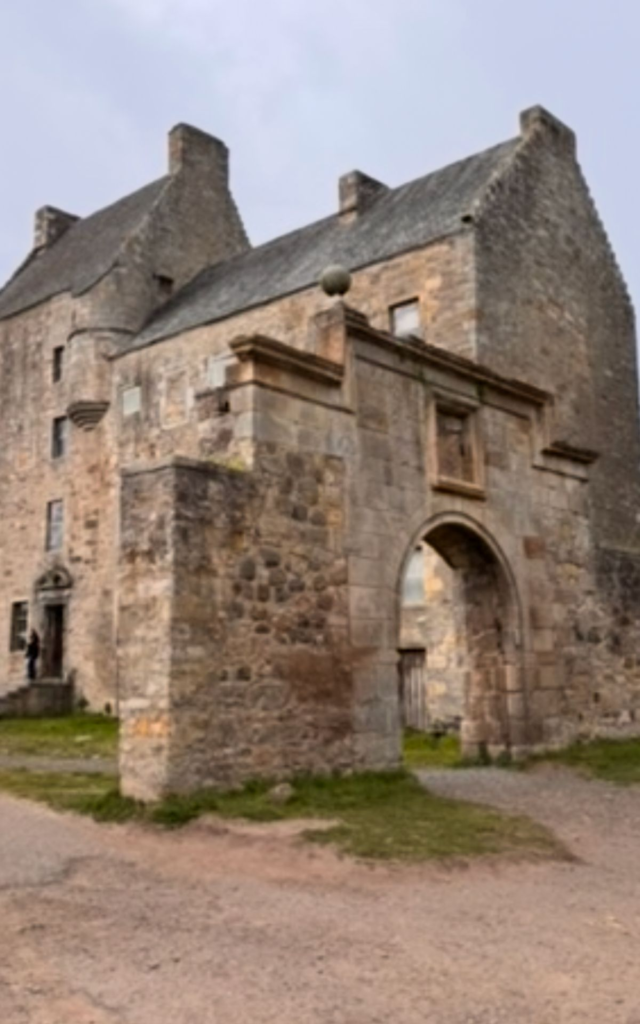 Stone castle with an arched entrance, pitched roofs, and weathered walls, set on a dirt path under a cloudy sky.