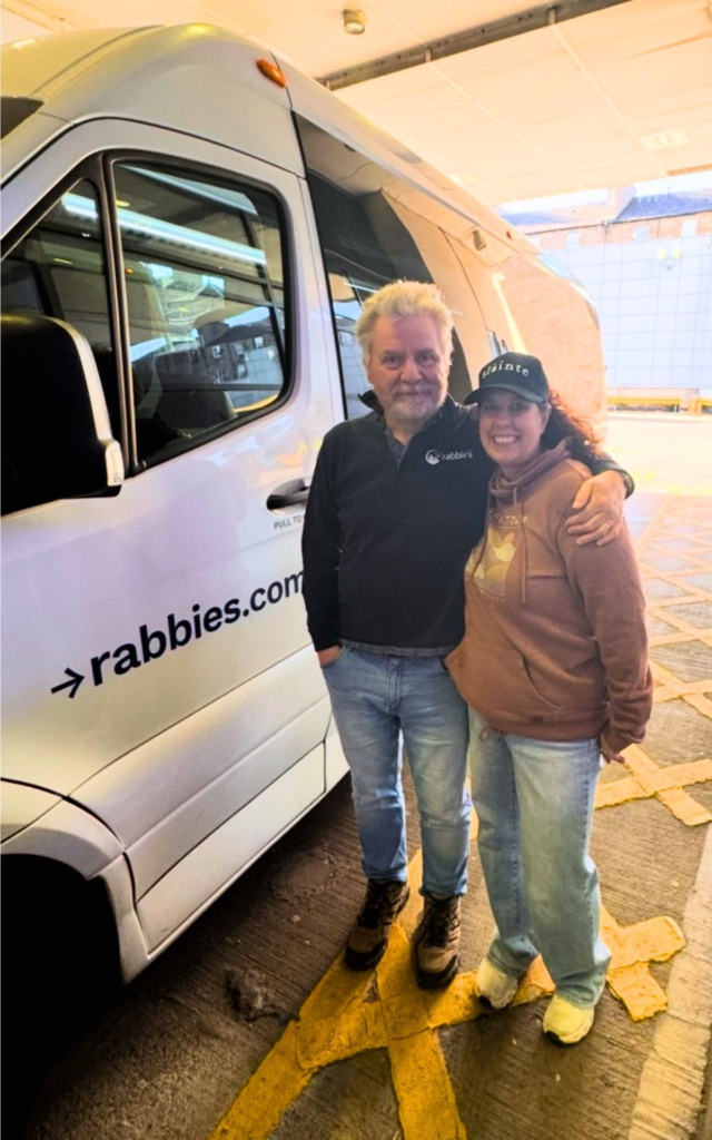 Two people stand smiling beside a white Rabbie’s tour van in a covered parking area. The man wears a dark jacket, and the woman wears a brown hoodie and a black cap.