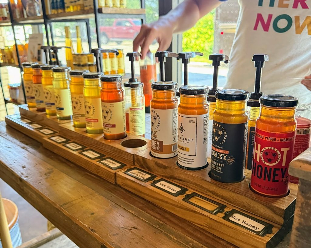 A person samples various honey flavors from labeled jars with pumps at a tasting station inside a store.