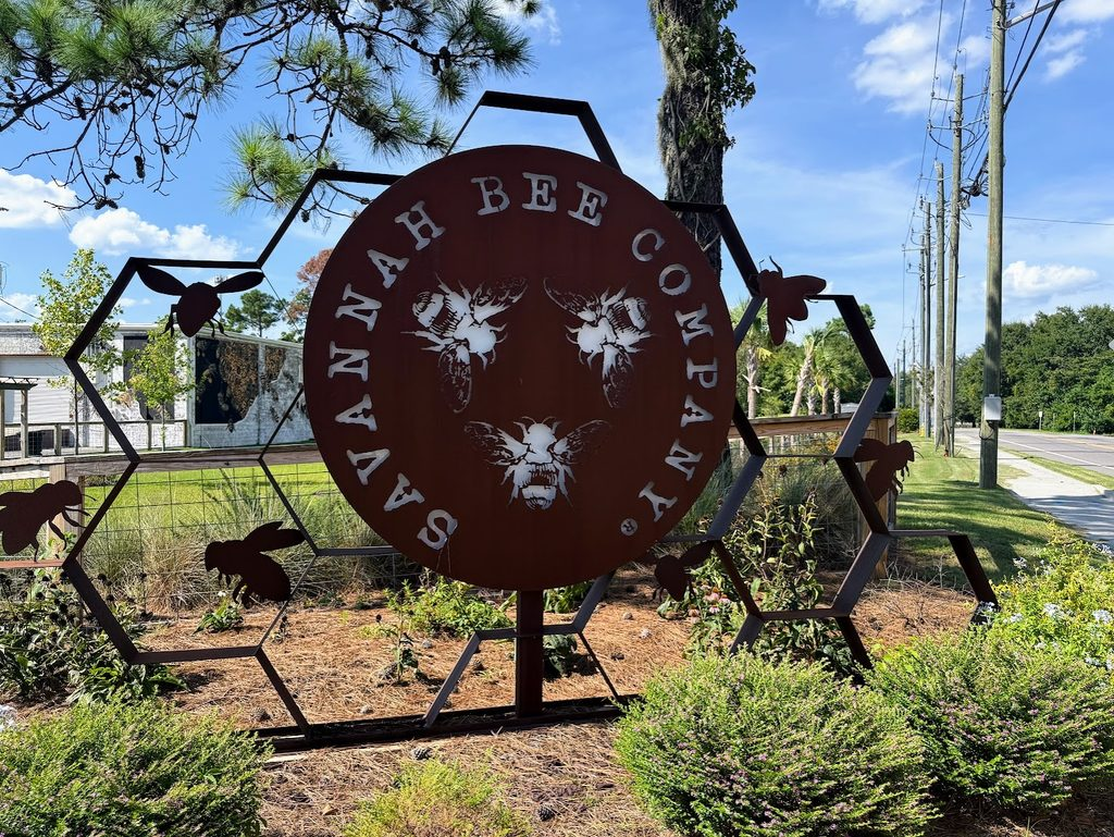 Large metal sign reads “Savannah Bee Company” with bee silhouettes, set among greenery by a roadside under a blue sky.