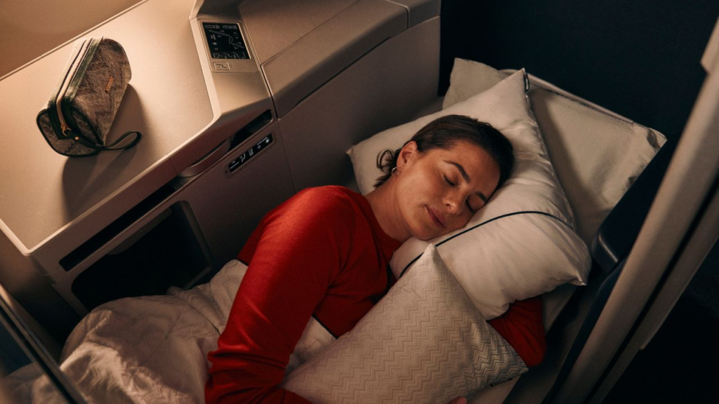 A woman in a red shirt sleeps in a lie-flat airline seat with an amenity kit on the side console.