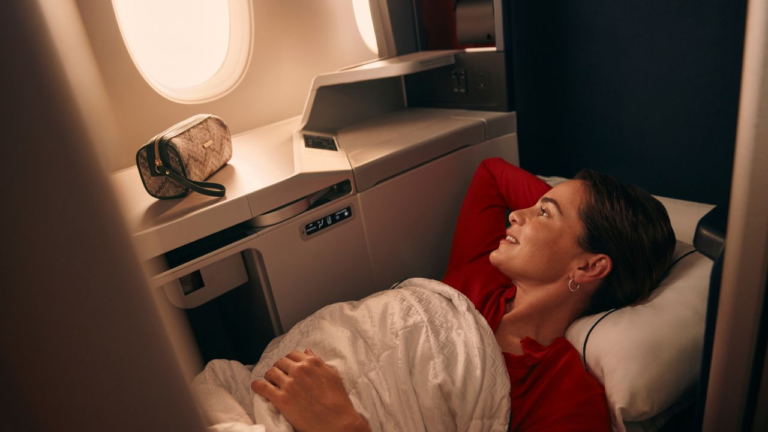 A woman in a red shirt reclines in a premium travel airplane bed with a blanket, looking toward a window next to a toiletry bag.