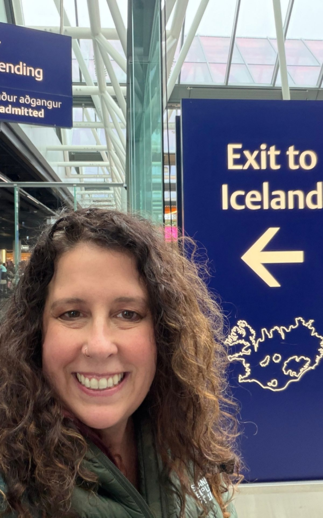 A smiling woman takes a selfie next to an airport sign that reads “Exit to Iceland” with an arrow and an outline map of the country.
