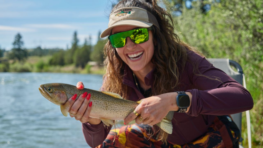 A woman wearing sunglasses and a visor holds a freshly caught trout by a river.