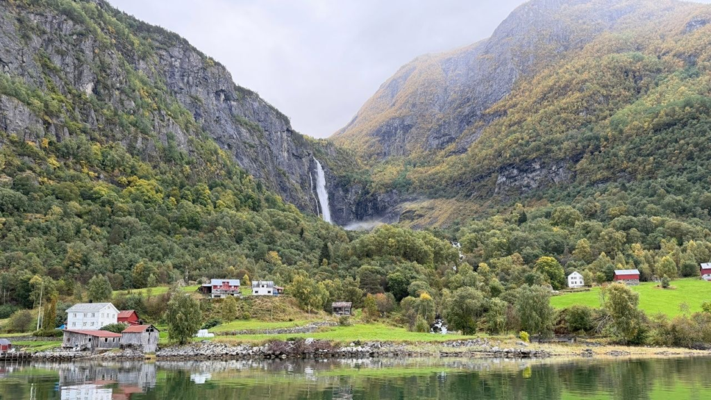A waterfall cascades down a steep mountain cliff above a small village with red and white houses on the shoreline of a fjord.