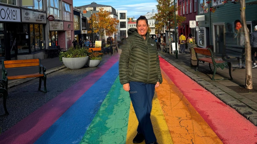 A smiling woman in an olive green jacket stands on a street painted with a rainbow stripe in front of city storefronts.